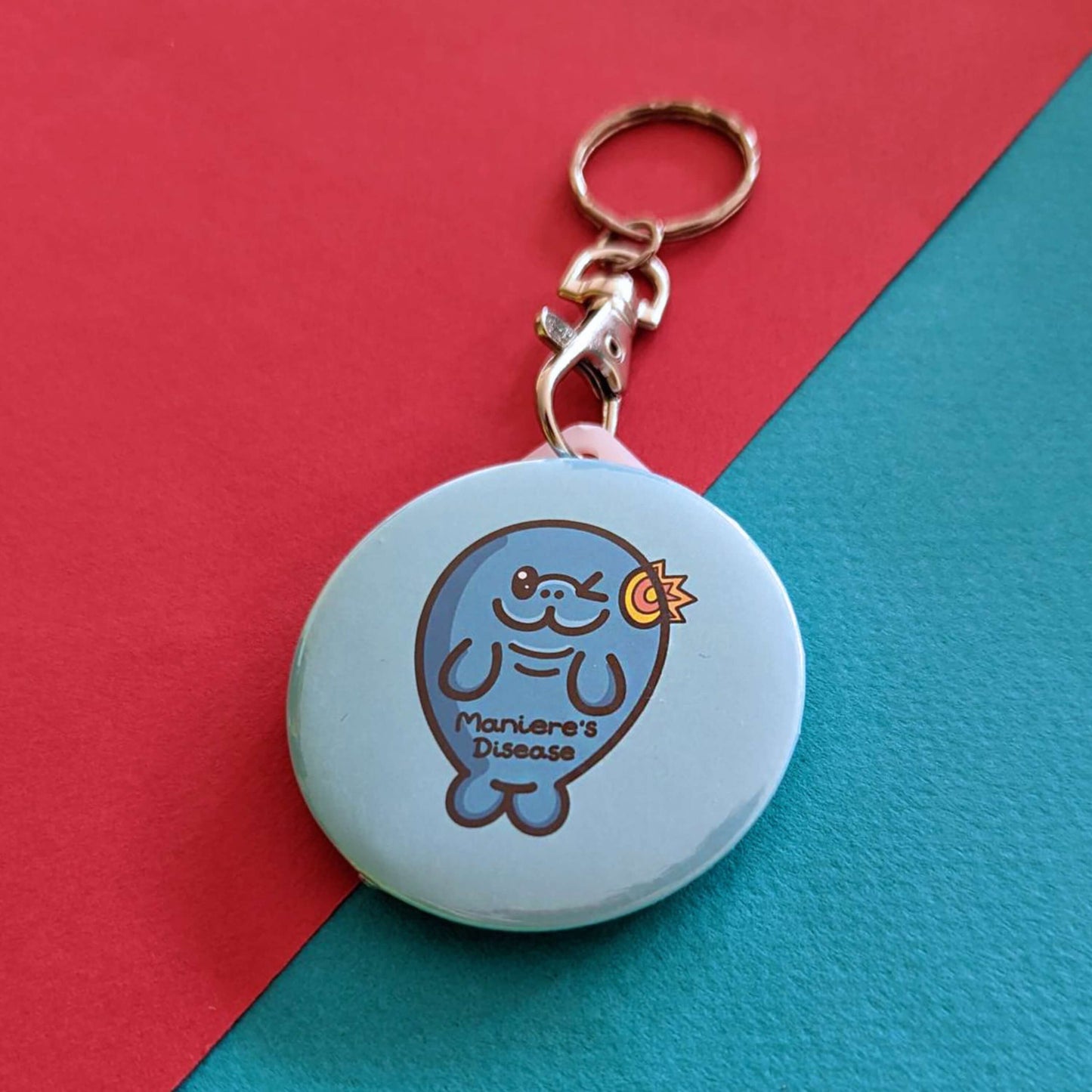 The Maniere's Disease Manatee Keyring - Ménière's Disease on a red and blue background. The pastel blue circular plastic keychain with silver lobster clip features a winking manatee with a yellow and red inflamed ear and black text across its body reading 'maniere's disease'. The hand drawn design is raising awareness for Ménière's Disease and vertigo.