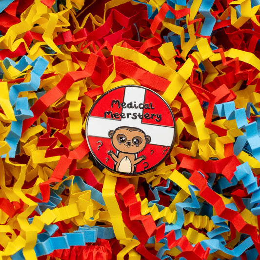 Medical Mystery Meerkat Enamel Pin on a red, blue and yellow crinkle card confetti background. The enamel pin is a red circle with white first aid cross and a cute meerkat at the bottom with big eyes and has it's little arms open wide. There are black question marks around the meerkat and 'medical meerstery' written above it in black writing. Hand drawn design made to raise awareness for invisible and chronic illnesses.