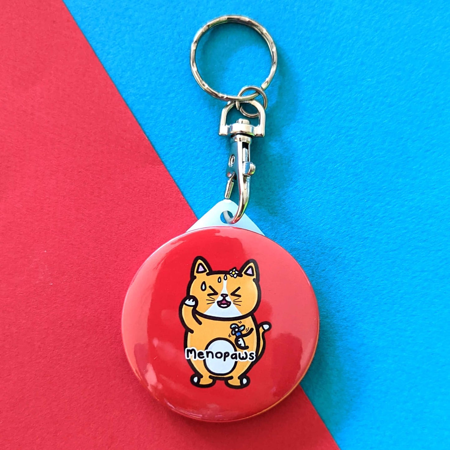 The Menopaws Cat Keyring - Menopause on a red and blue background. The red circular keychain features a stressed sweating orange cat standing up holding a blue mini fan, underneath in black text reading 'menopaws'. Raising awareness for the menopause and menopausal symptoms.