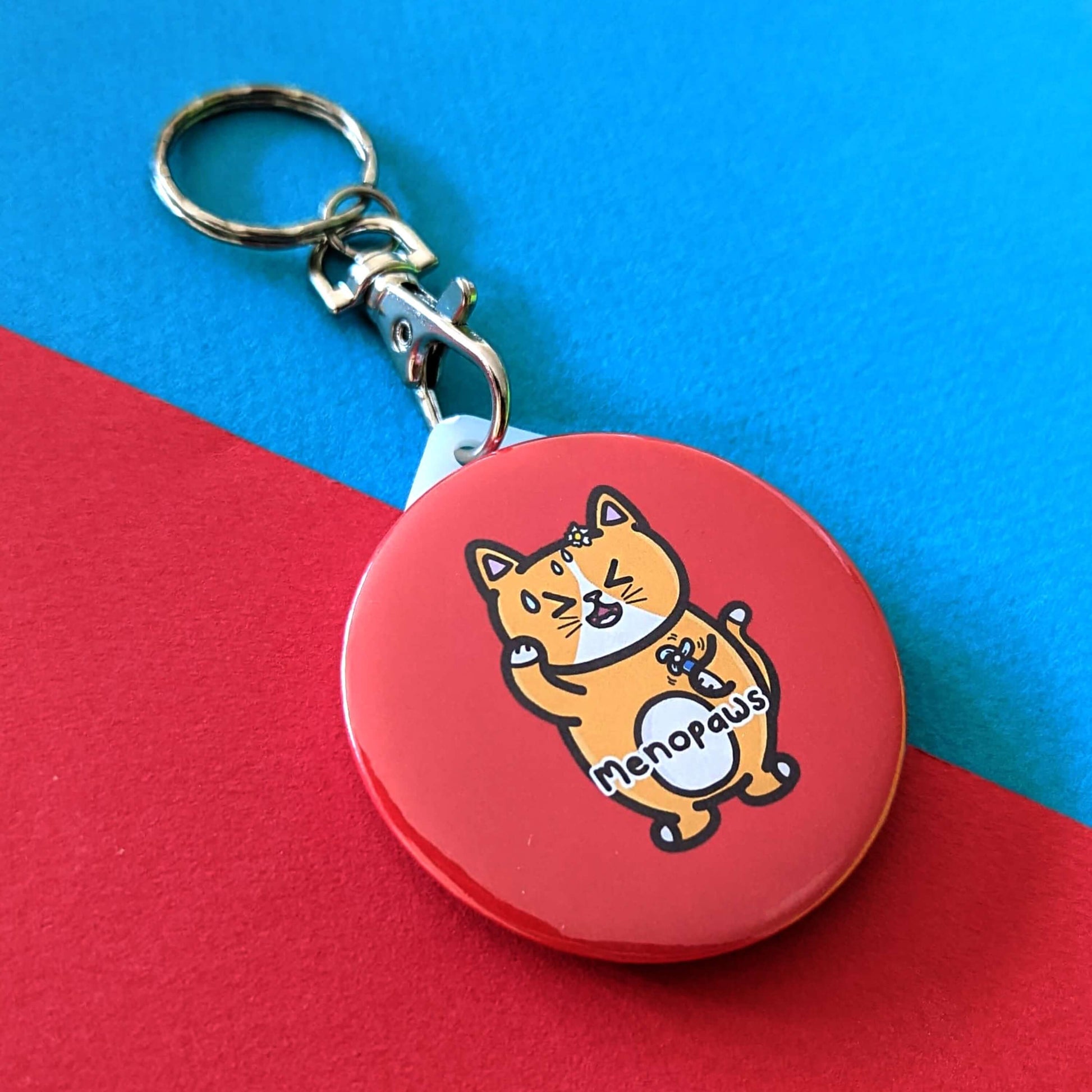 The Menopaws Cat Keyring - Menopause on a red and blue background. The red circular keychain features a stressed sweating orange cat standing up holding a blue mini fan, underneath in black text reading 'menopaws'. Raising awareness for the menopause and menopausal symptoms.