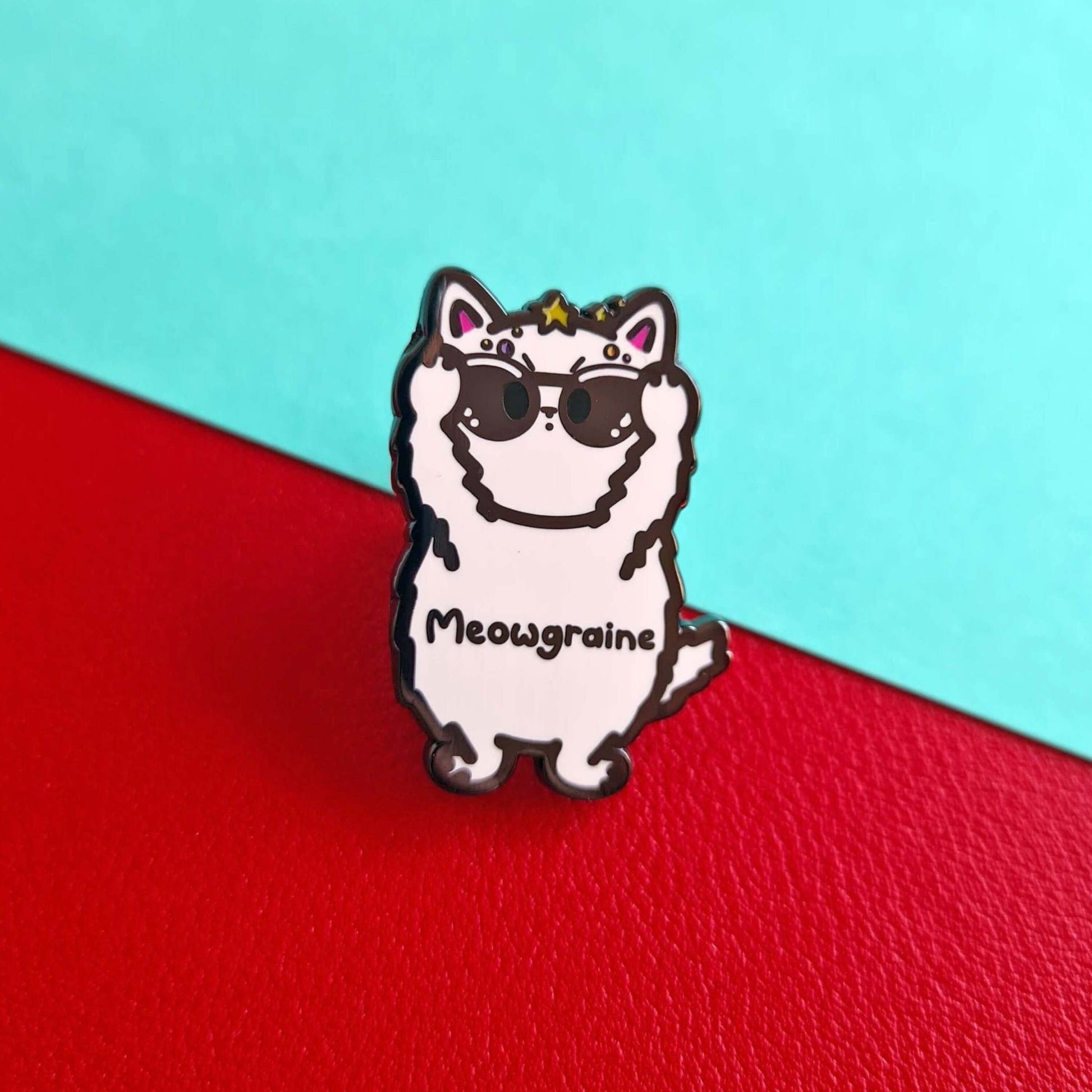 The Meowgraine Cat Enamel Pin - Migraine on a red and blue background. A white stressed cat clutching a pair of black sunglasses to its eyes with multicoloured spots and stars over its head, across its middle reads 'meowgraine'. The hand drawn design is raising awareness for migraines and headaches.