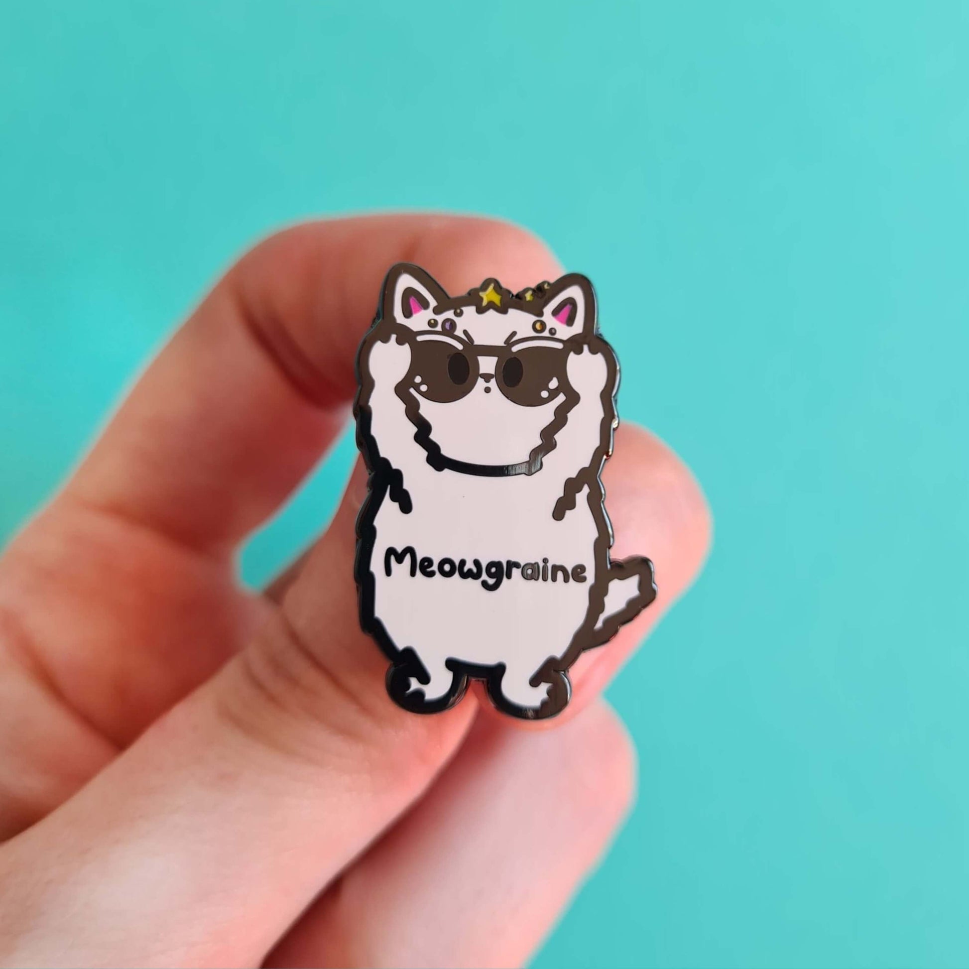 The Meowgraine Cat Enamel Pin - Migraine held over a blue background. A white stressed cat clutching a pair of black sunglasses to its eyes with multicoloured spots and stars over its head, across its middle reads 'meowgraine'. The hand drawn design is raising awareness for migraines and headaches.
