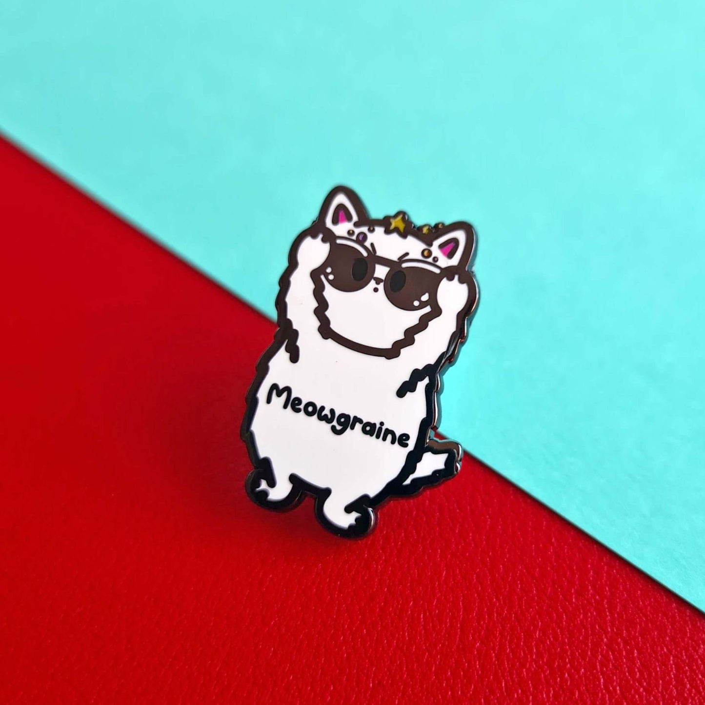 The Meowgraine Cat Enamel Pin - Migraine on a red and blue background. A white stressed cat clutching a pair of black sunglasses to its eyes with multicoloured spots and stars over its head, across its middle reads 'meowgraine'. The hand drawn design is raising awareness for migraines and headaches.