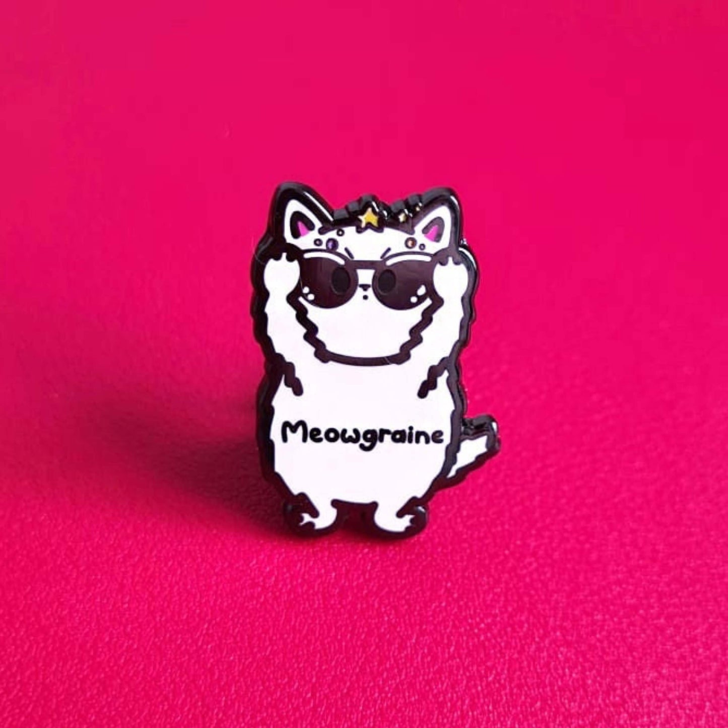 The Meowgraine Cat Enamel Pin - Migraine on a red background. A white stressed cat clutching a pair of black sunglasses to its eyes with multicoloured spots and stars over its head, across its middle reads 'meowgraine'. The hand drawn design is raising awareness for migraines and headaches.