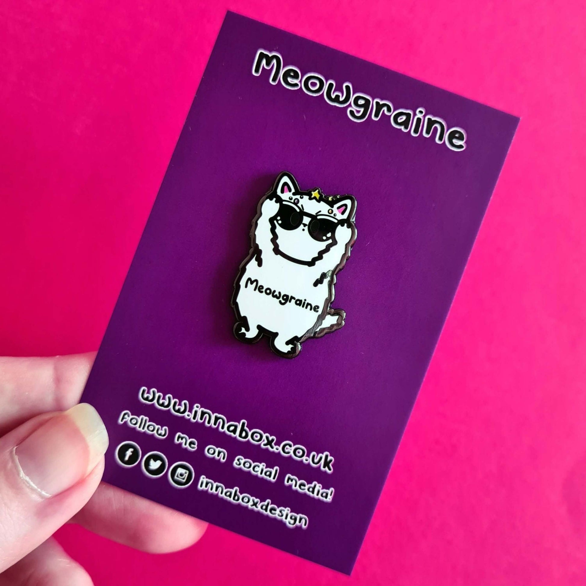 The Meowgraine Cat Enamel Pin - Migraine on purple backing card held over a red background. A white stressed cat clutching a pair of black sunglasses to its eyes with multicoloured spots and stars over its head, across its middle reads 'meowgraine'. The hand drawn design is raising awareness for migraines and headaches.