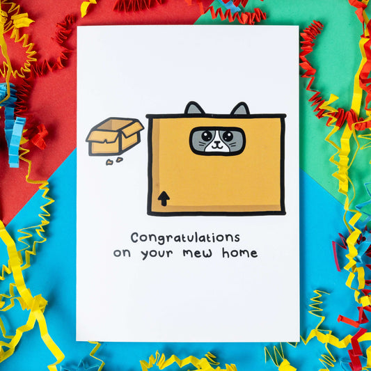 The Mew Home Cat Card on a red, blue and green background with red, yellow and blue crinkle card confetti. The white a6 greeting card features two brown moving boxes, one with a smiling grey cat hiding inside. Underneath in black text reads 'congratulations on your mew home'. 
