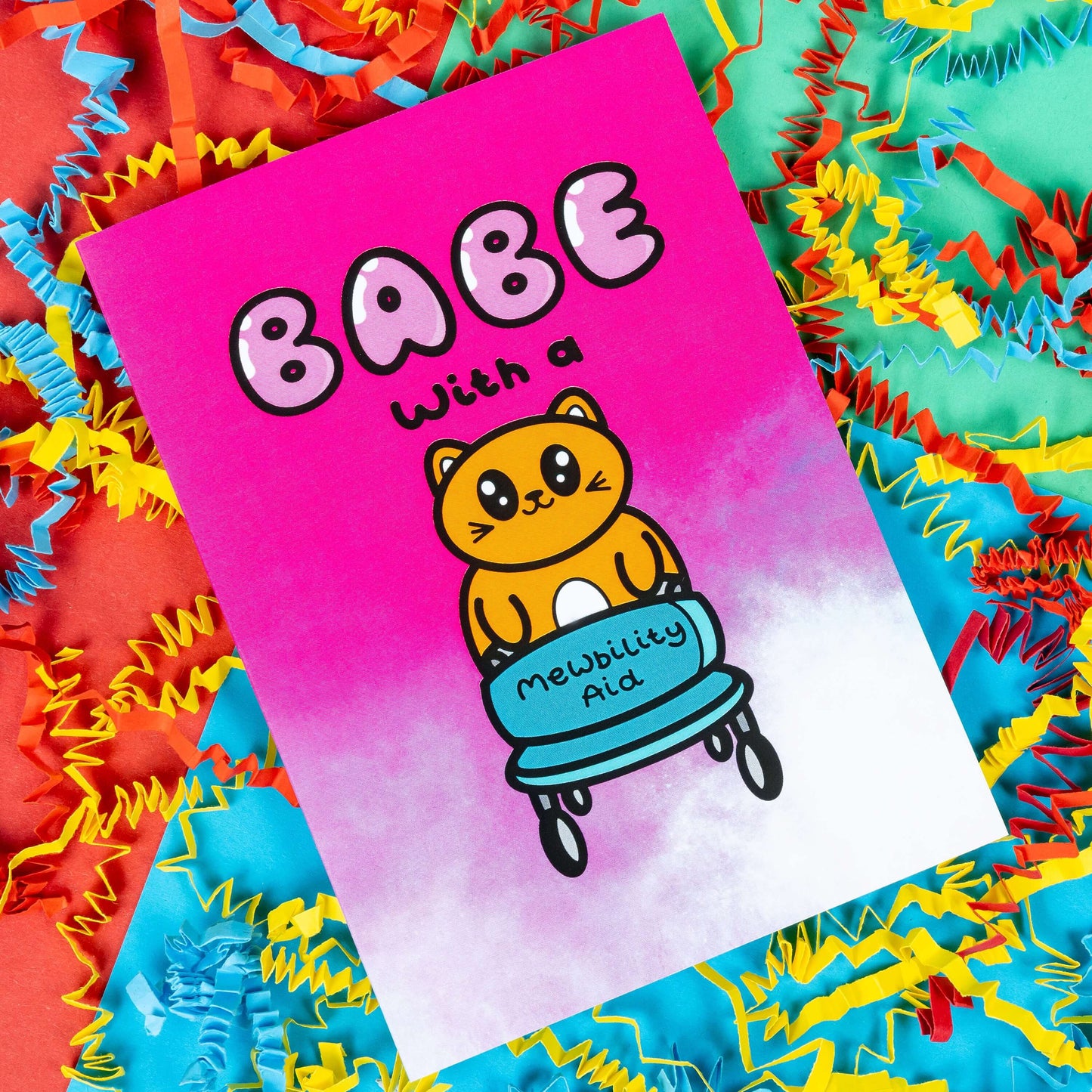 A hot pink and white gradient greeting card with a cute ginger cat holding a blue mobility aid. 'Babe with a mewbility aid' is written on the card. The background of the photo is colourful card confetti. Design inspired by disabilities and chronic illness.