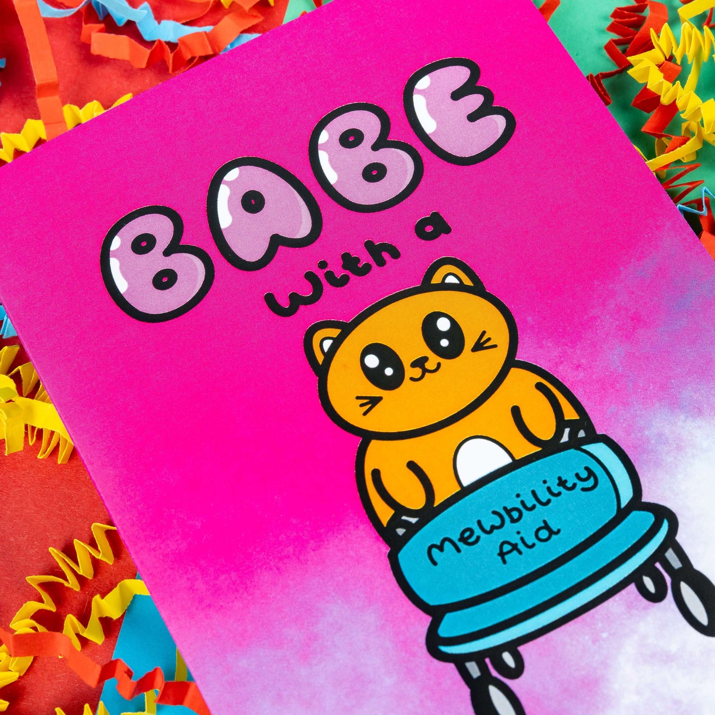 Close up of a hot pink and white gradient greeting card with a cute ginger cat holding a blue mobility aid. 'Babe with a mewbility aid' is written on the card. The background of the photo is colourful card confetti. Design inspired by disabilities and chronic illness.