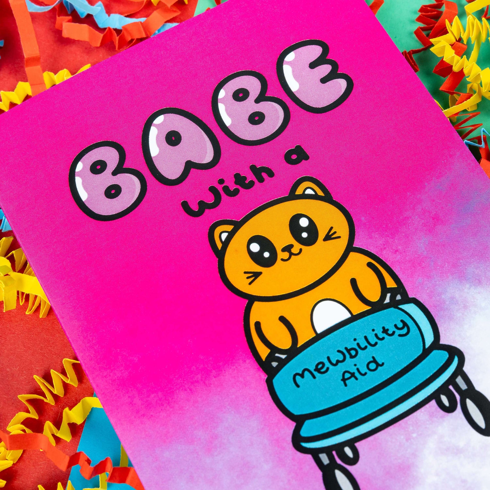 Close up of a hot pink and white gradient greeting card with a cute ginger cat holding a blue mobility aid. 'Babe with a mewbility aid' is written on the card. The background of the photo is colourful card confetti. Design inspired by disabilities and chronic illness.