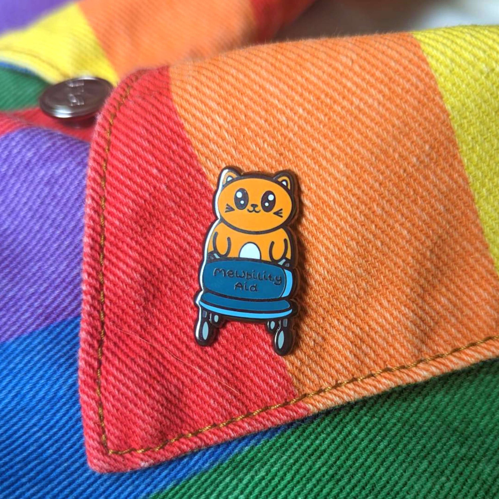 The Mewbility Aid Cat Enamel Pin - Mobility Aid on a rainbow denim jacket. An orange smiling cat walking upright with a blue mobility aid, across the walking chair reads 'mewbility aid'. The hand drawn designs is raising awareness for mobility aids.