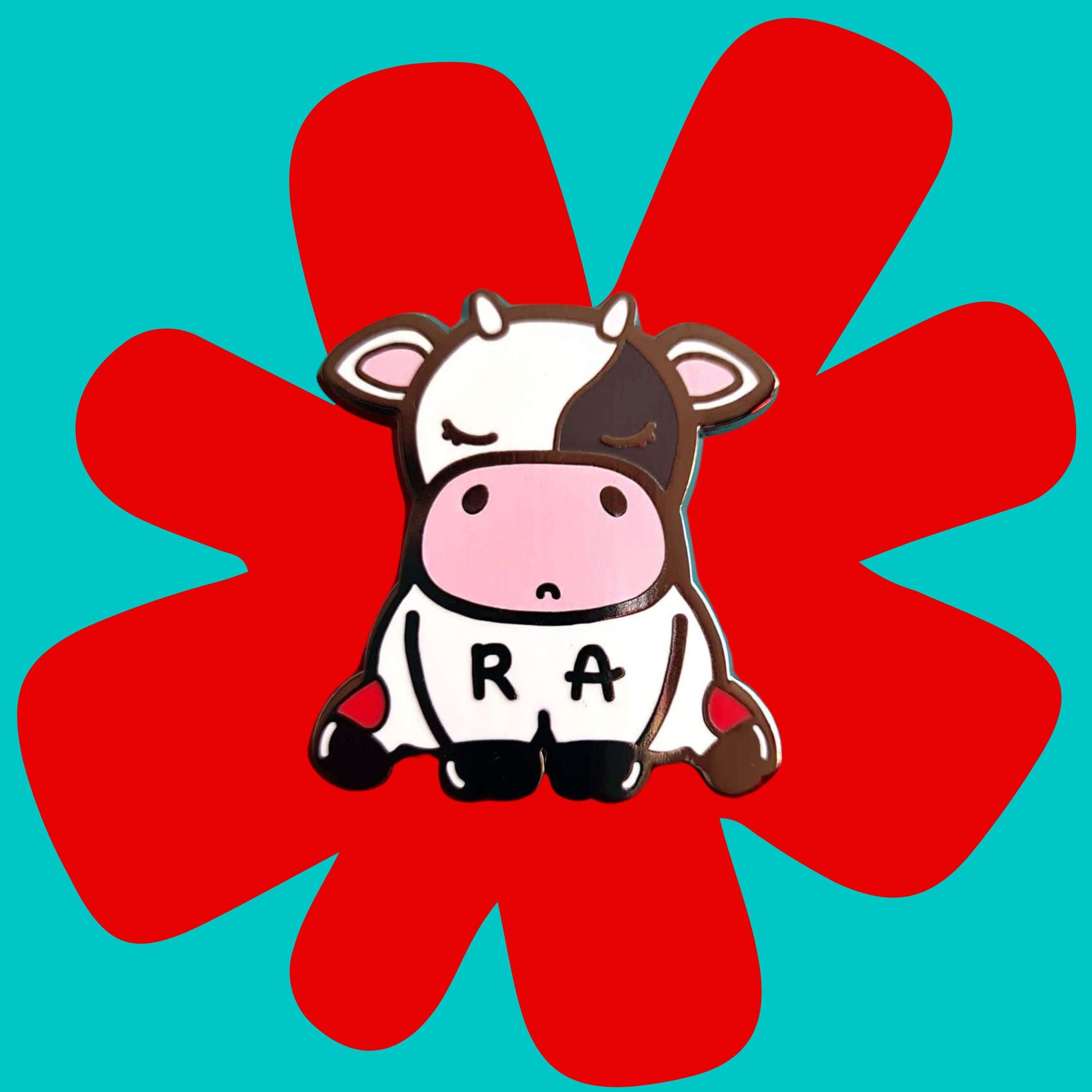 The Moomatoid Arthritis Cow Enamel Pin - Rheumatoid Arthritis on a red and blue background. The sad cow shape pin has red marks on its lower back legs and the initials R and A across its middle. The hand drawn design is raising awareness for Rheumatoid Arthritis.