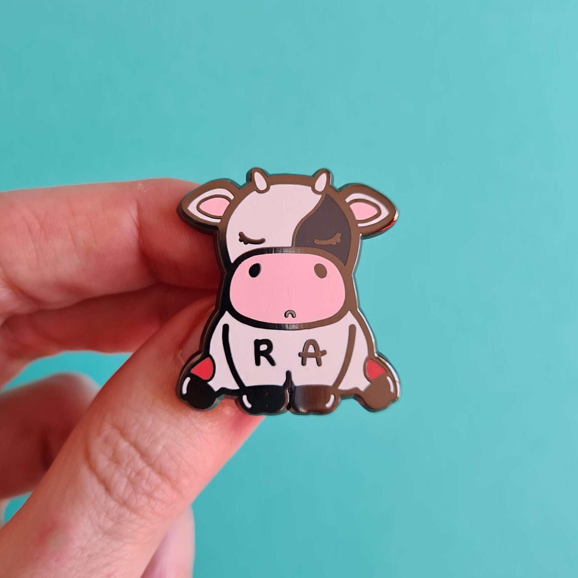 The Moomatoid Arthritis Cow Enamel Pin - Rheumatoid Arthritis held over a blue background. The sad cow shape pin has red marks on its lower back legs and the initials R and A across its middle. The hand drawn design is raising awareness for Rheumatoid Arthritis.