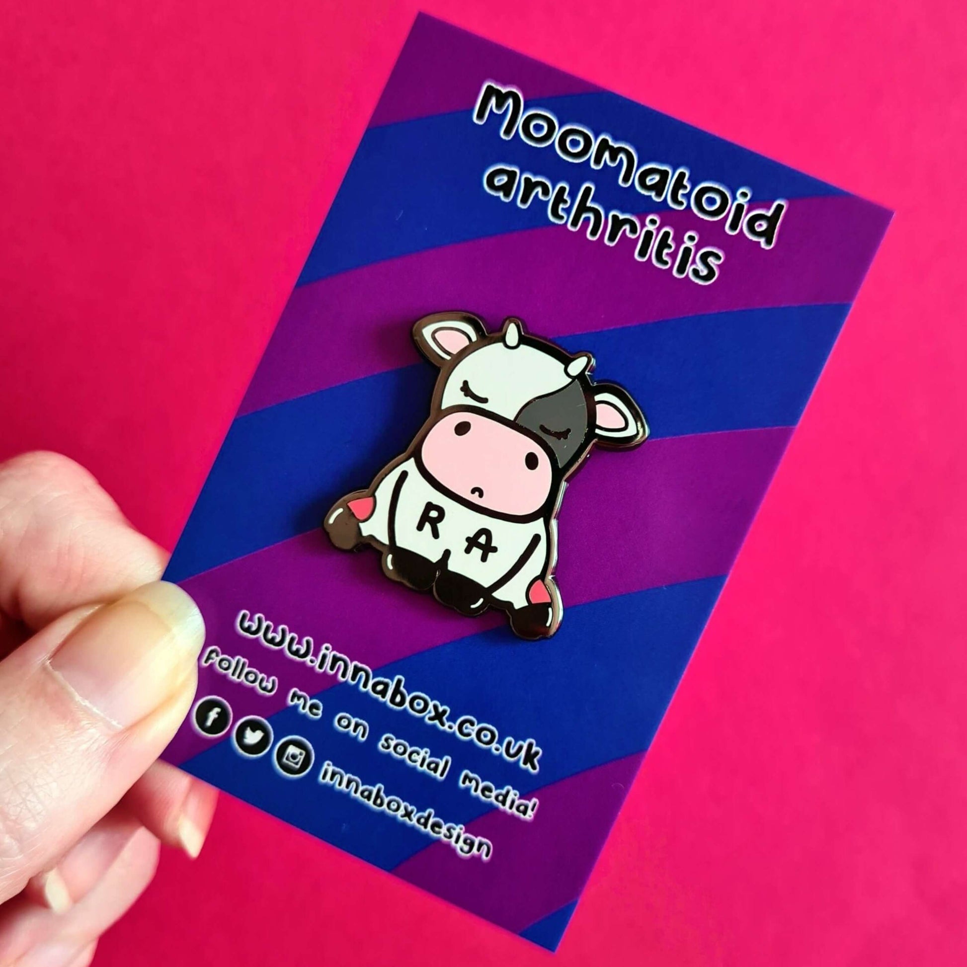 The Moomatoid Arthritis Cow Enamel Pin - Rheumatoid Arthritis on purple and blue stripe backing card held over a red background. The sad cow shape pin has red marks on its lower back legs and the initials R and A across its middle. The hand drawn design is raising awareness for Rheumatoid Arthritis.
