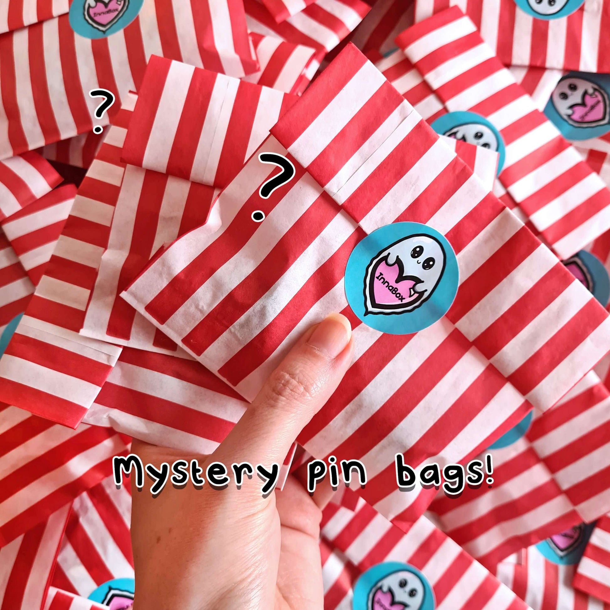 The Mystery Enamel Pin Bag held over a pile of the mystery bags. They are red and white stripe candy bags sealed with the innabox blue ghost logo sticker with black text at the bottom reading 'mystery pin bags!'. A surprise goodie bag full of three enamel pins raising awareness for hidden disabilities and invisible illnesses.