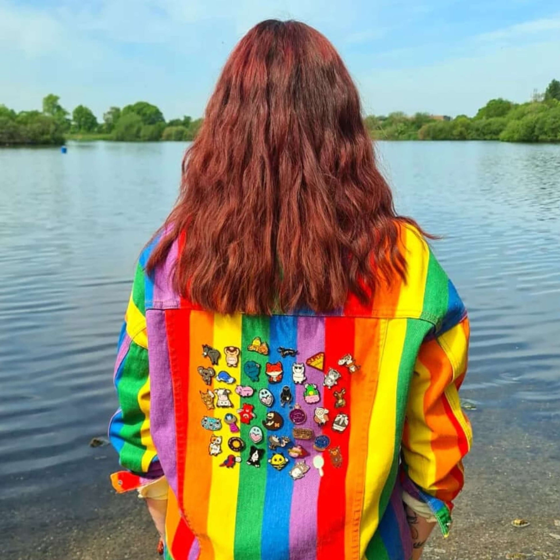 Nikky, the owner of Innabox, with brown mid length hair facing away from the camera looking out to a lake. She is wearing a rainbow denim jacket with the back full of the innabox enamel pins raising awareness for hidden disabilities and invisible illnesses.