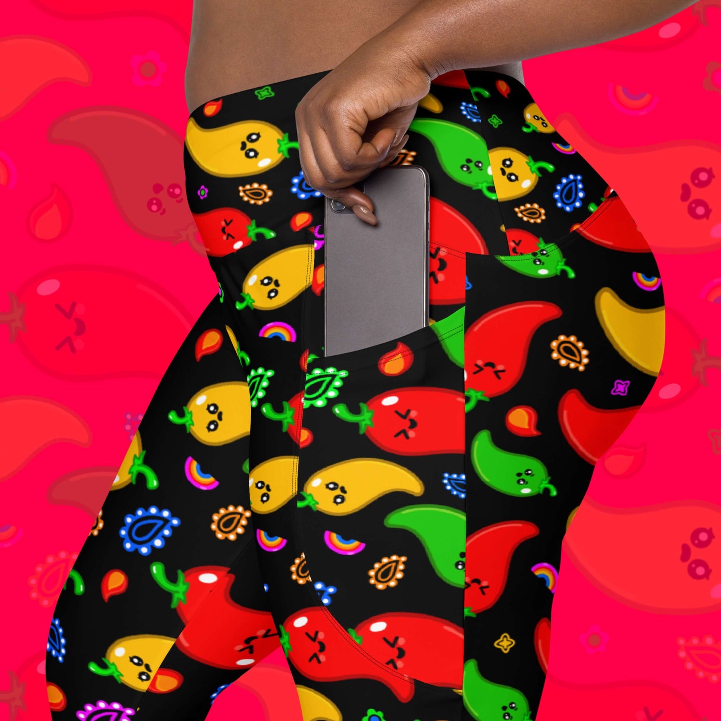 Black leggings with red, green, orange and yellow chilli peppers with cute faces on and various cute doodles around them shown on model who is removing a phone from the pocket of the leggings. The background of the photo is a faded close up of the print. The hand drawn design is raising awareness for neurodiversity. 