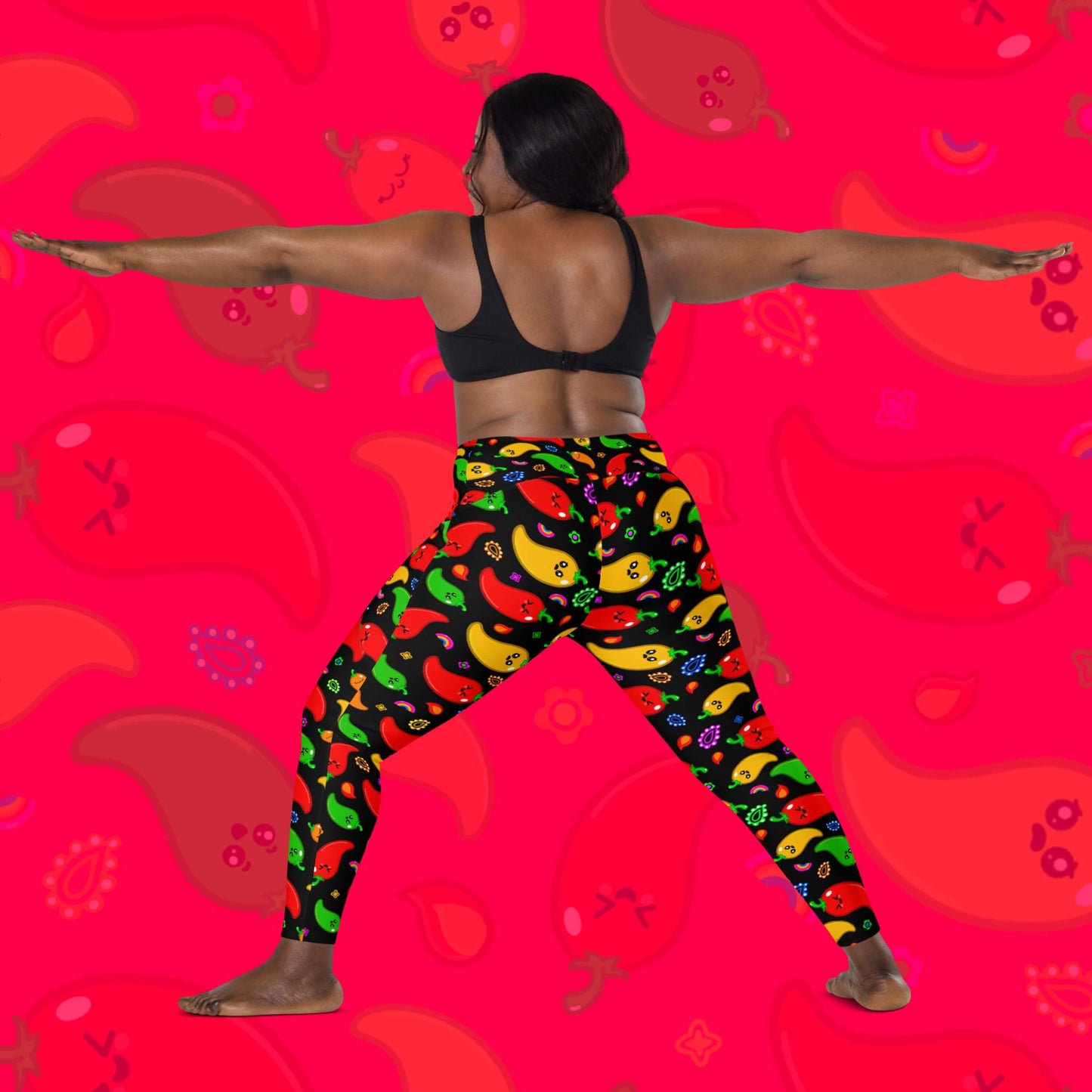 Black leggings with pockets with red, green, orange and yellow chilli peppers with cute faces on and various cute doodles around them shown on a model who is doing a yoga pose. The background of the photo is a faded close up of the print. The hand drawn design is raising awareness for neurodiversity. 