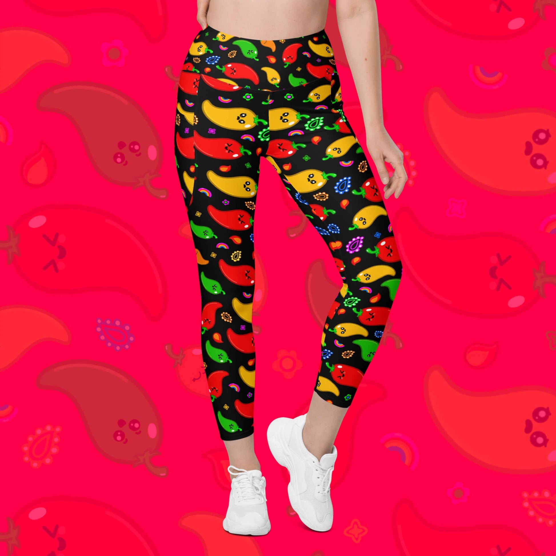Black leggings with pockets with red, green, orange and yellow chilli peppers with cute faces on and various cute doodles around them shown on a model who has one leg bent and the other straight. The background of the photo is a faded close up of the print. The hand drawn design is raising awareness for neurodiversity. 