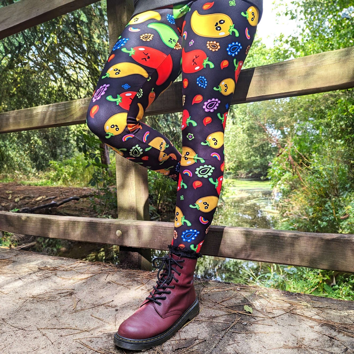A person leaning against a fence wearing Black leggings with red, green, orange and yellow chilli peppers with cute faces on and various cute doodles around them. The hand drawn design is raising awareness for neurodiversity.