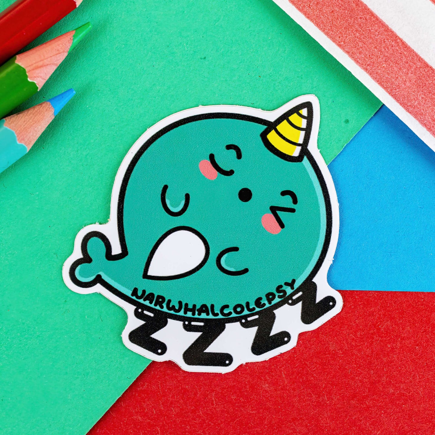 The Narwhalcolepsy Narwhal Sticker - Narcolepsy on a red, blue and green background with colouring pencils and red stripe candy bag. The blue green narwhal shape sticker is sleeping on 4 black Z's with black text reading 'narhwalcolepsy' across its body. The hand drawn design is raising awareness for narcolepsy.