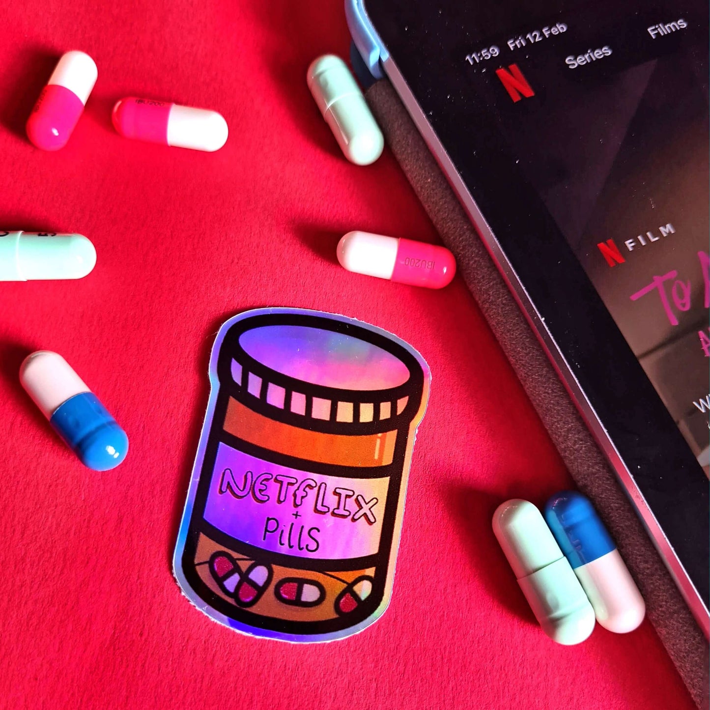 The Netflix & Pills Holographic Sticker on a red background with multicoloured prescription pills and a phone with the netflix app open. The holographic shiny rainbow pill bottle sticker filled with red and white tablets with a label that reads 'netflix + pills'. A cheeky hand drawn design to raise awareness for hidden disability prescriptions.