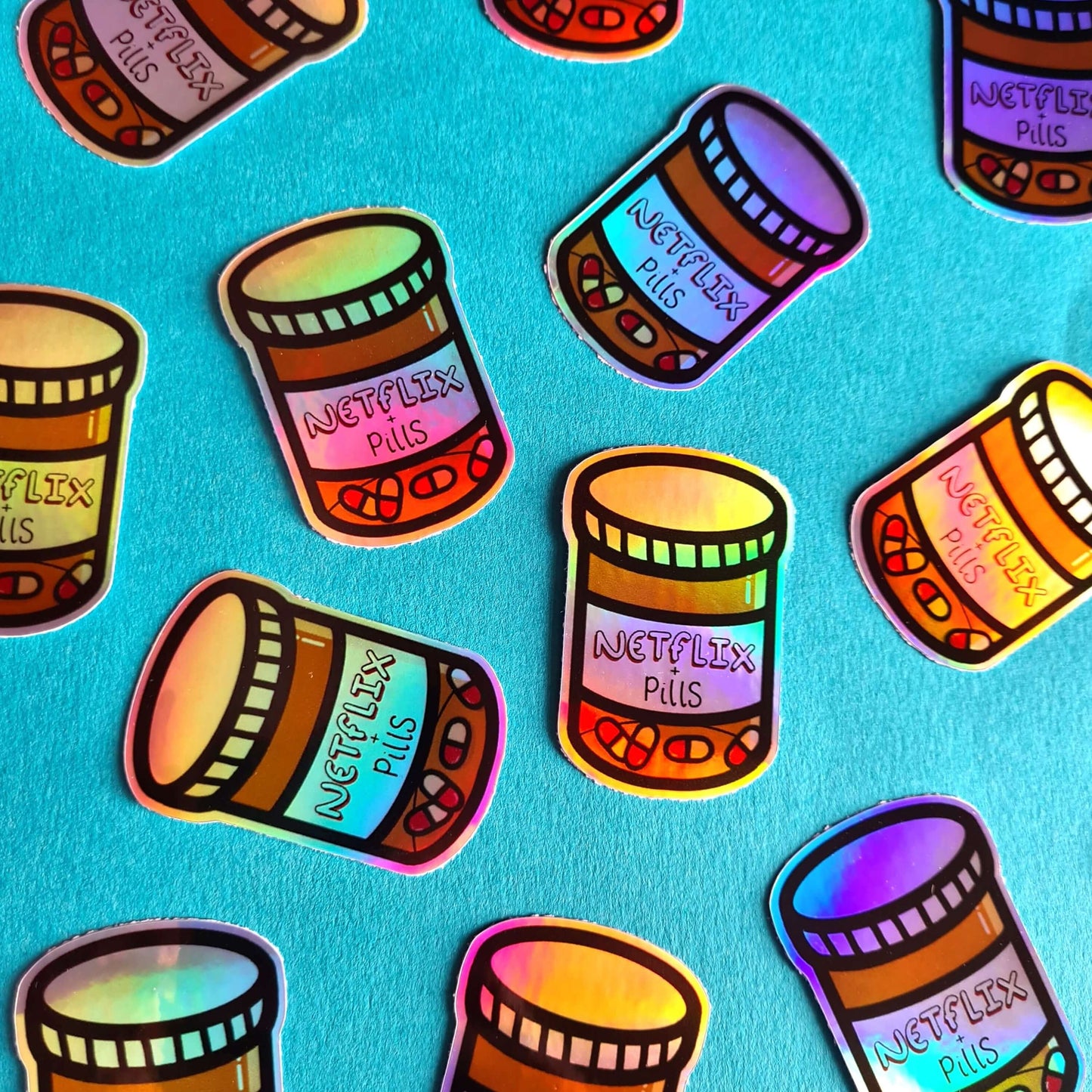 The Netflix & Pills Holographic Sticker on a blue background with multiple stickers of the same design. The holographic shiny rainbow pill bottle sticker filled with red and white tablets with a label that reads 'netflix + pills'. A cheeky hand drawn design to raise awareness for hidden disability prescriptions.