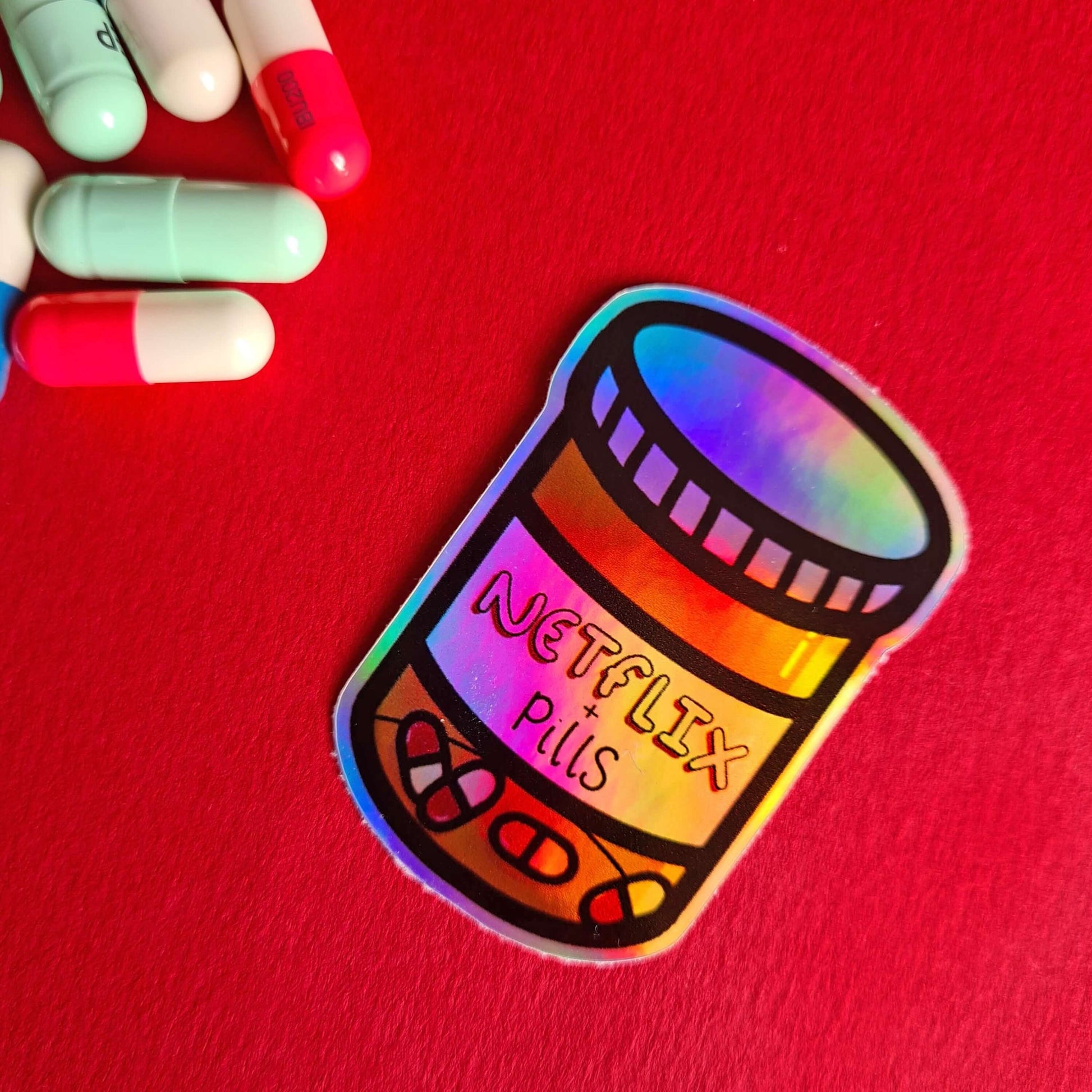 The Netflix & Pills Holographic Sticker on a red background with multicoloured tablets. The holographic shiny rainbow pill bottle sticker filled with red and white tablets with a label that reads 'netflix + pills'. A cheeky hand drawn design to raise awareness for hidden disability prescriptions.