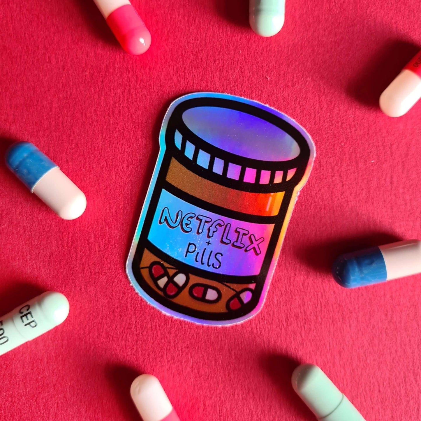 The Netflix & Pills Holographic Sticker on a red background with multicoloured tablets. The holographic shiny rainbow pill bottle sticker filled with red and white tablets with a label that reads 'netflix + pills'. A cheeky hand drawn design to raise awareness for hidden disability prescriptions.
