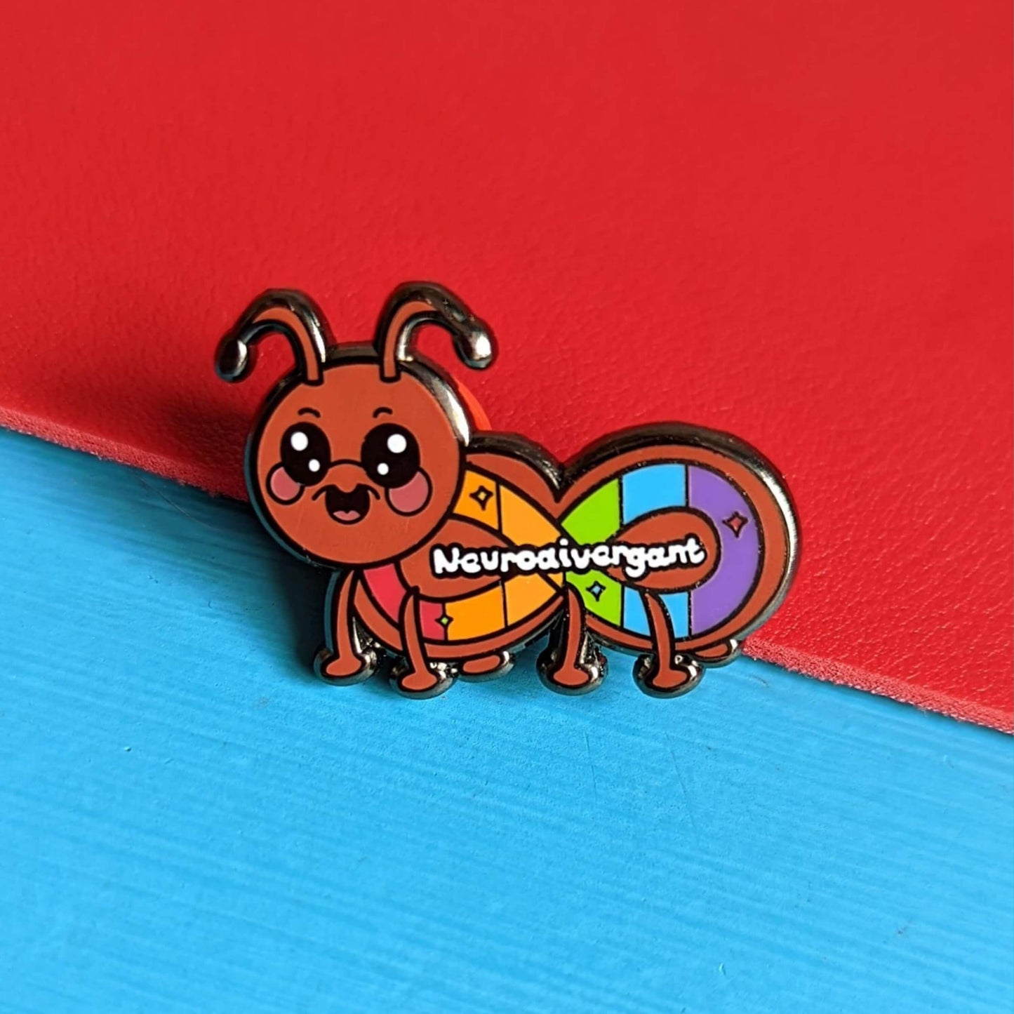 The Neurodivergant Ant Enamel Pin - Neurodivergent on a red and blue background. The brown ant shape enamel pin is smiling with a rainbow infinity symbol and white text reading 'neurodivergant' with sparkles across its body. The hand drawn design is raising awareness for neurodiversity.