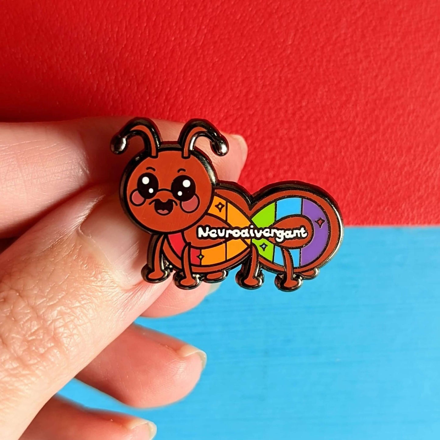 The Neurodivergant Ant Enamel Pin - Neurodivergent on a red and blue background. The brown ant shape enamel pin is smiling with a rainbow infinity symbol and white text reading 'neurodivergant' with sparkles across its body. The hand drawn design is raising awareness for neurodiversity.