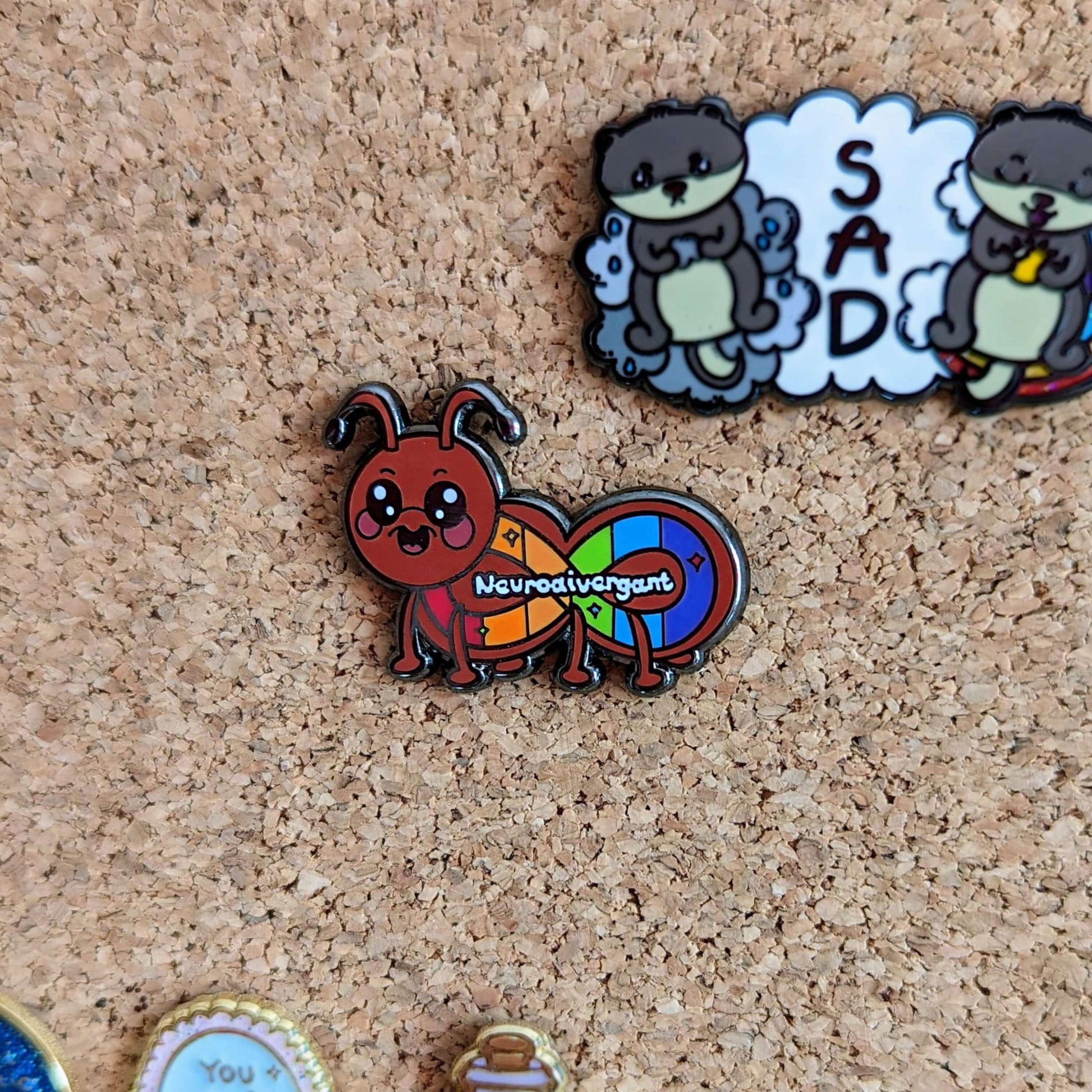 The Neurodivergant Ant Enamel Pin - Neurodivergent pinned on a cork pin board. The brown ant shape enamel pin is smiling with a rainbow infinity symbol and white text reading 'neurodivergant' with sparkles across its body. The hand drawn design is raising awareness for neurodiversity.