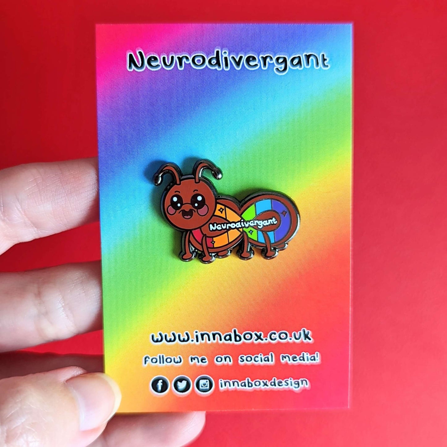 The Neurodivergant Ant Enamel Pin - Neurodivergent on rainbow backing card with innabox social media handles along the bottom held over a red background. The brown ant shape enamel pin is smiling with a rainbow infinity symbol and white text reading 'neurodivergant' with sparkles across its body. The hand drawn design is raising awareness for neurodiversity.