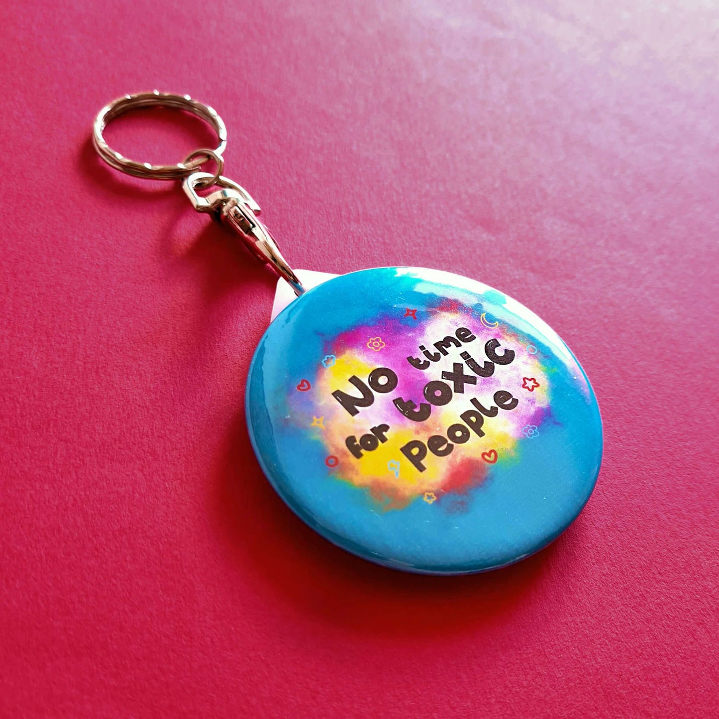 The No Time For Toxic People Keyring on a red background. The silver lobster clip blue plastic circular keychain has yellow and pink clouds with black text reading 'no time for toxic people' with red, yellow, and blue hearts, clouds, sparkles, moons and flowers.