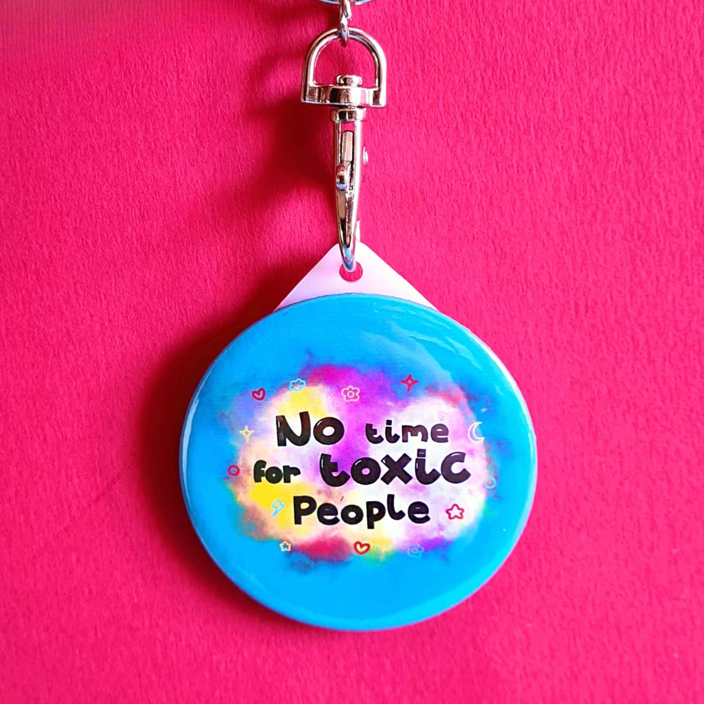 The No Time For Toxic People Keyring on a red background. The silver lobster clip blue plastic circular keychain has yellow and pink clouds with black text reading 'no time for toxic people' with red, yellow, and blue hearts, clouds, sparkles, moons and flowers.