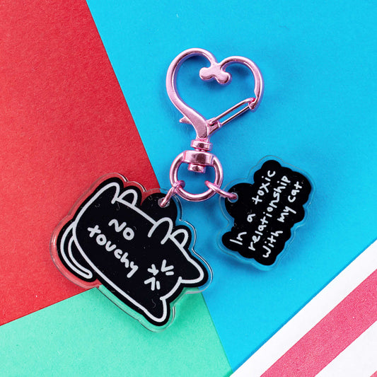 The No Touchy Cat Keyring on a red, blue and green card background. The pink metal heart clip keyring has two acrylic charms, a black frustrated cat with white text reading 'no touchy' and a black cloud with white text reading 'in a toxic relationship with my cat'.