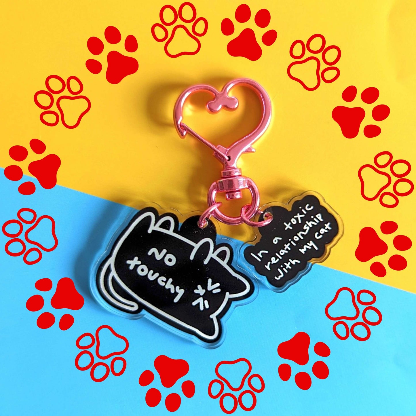 The No Touchy Cat Keyring on a blue and yellow card background with red paw prints. The pink metal heart clip keyring has two acrylic charms, a black frustrated cat with white text reading 'no touchy' and a black cloud with white text reading 'in a toxic relationship with my cat'.