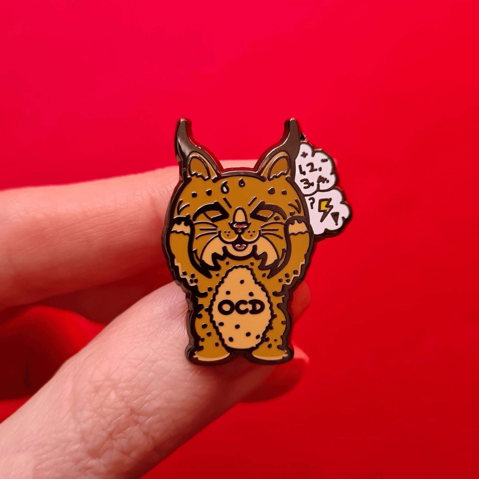 Bobsessive Compulsive Disorder Enamel Pin - OCD being held over a red background. The bobcat shape pin is brown with a stressed expression clutching its face with a thought bubble of numbers, question marks and lightning bolts. Across its tummy reads OCD. The pin was designed to raise awareness of obsessive compulsive disorder.