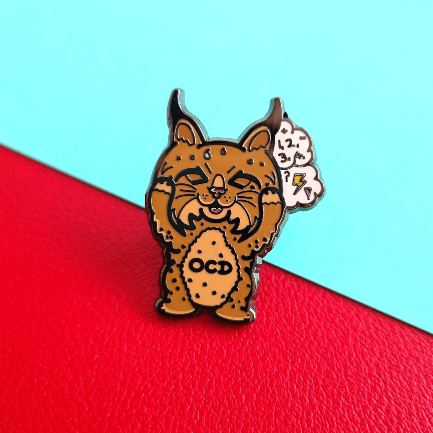 Bobsessive Compulsive Disorder Enamel Pin - OCD on a red and blue background. The bobcat shape pin is brown with a stressed expression clutching its face with a thought bubble of numbers, question marks and lightning bolts. Across its tummy reads OCD. The pin was designed to raise awareness of obsessive compulsive disorder.