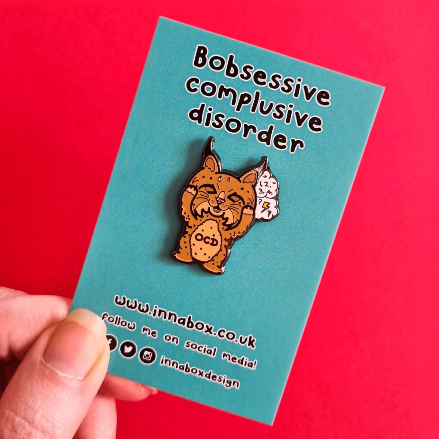 Bobsessive Compulsive Disorder Enamel Pin - OCD on blue backing card with innabox social media info being held over a red background. The bobcat shape pin is brown with a stressed expression clutching its face with a thought bubble of numbers, question marks and lightning bolts. Across its tummy reads OCD. The pin was designed to raise awareness of obsessive compulsive disorder.