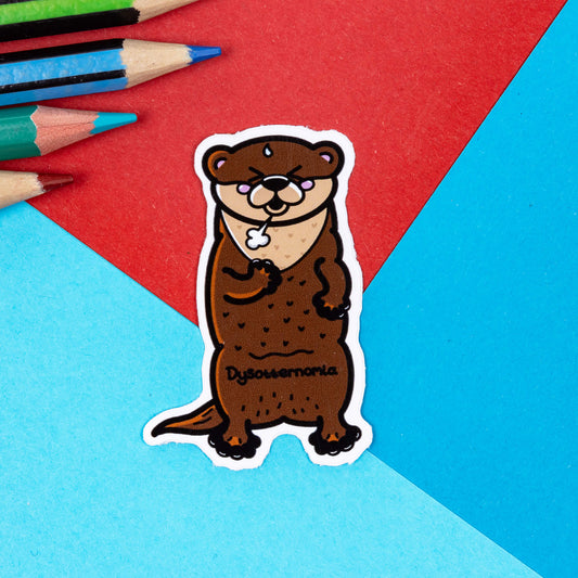 The Dysotternomia Otter Sticker - Dysautonomia on a red and blue background with colouring pencils. The brown otter sticker is wheezing clutching its chest with black text across its body reading 'dysotternomia'. The hand drawn design is raising awareness for Dysautonomia.