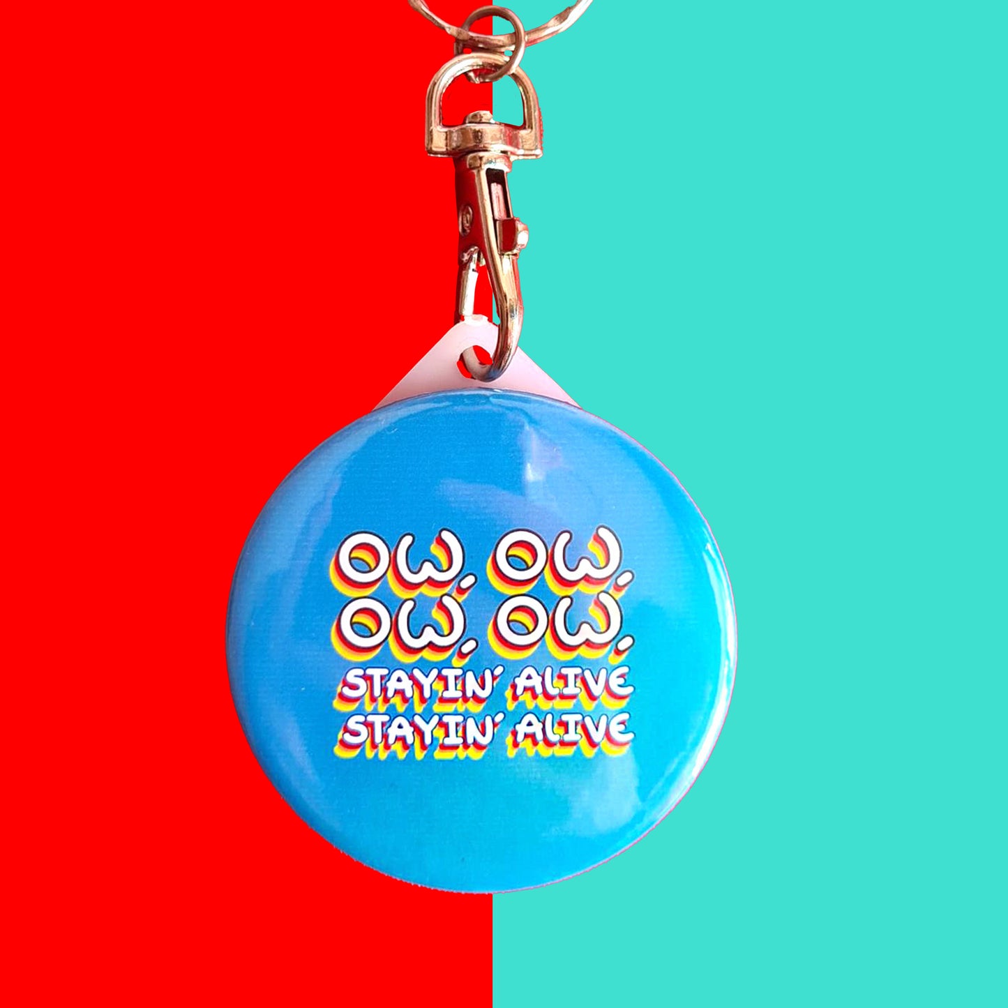 The Ow, Ow, Ow, Ow, Staying Alive Keyring on a blue and red background. The silver lobster clip plastic blue circular keychain with white text with red, orange and yellow borders reading 'ow, ow, ow, ow, stayin' alive stayin' alive'.