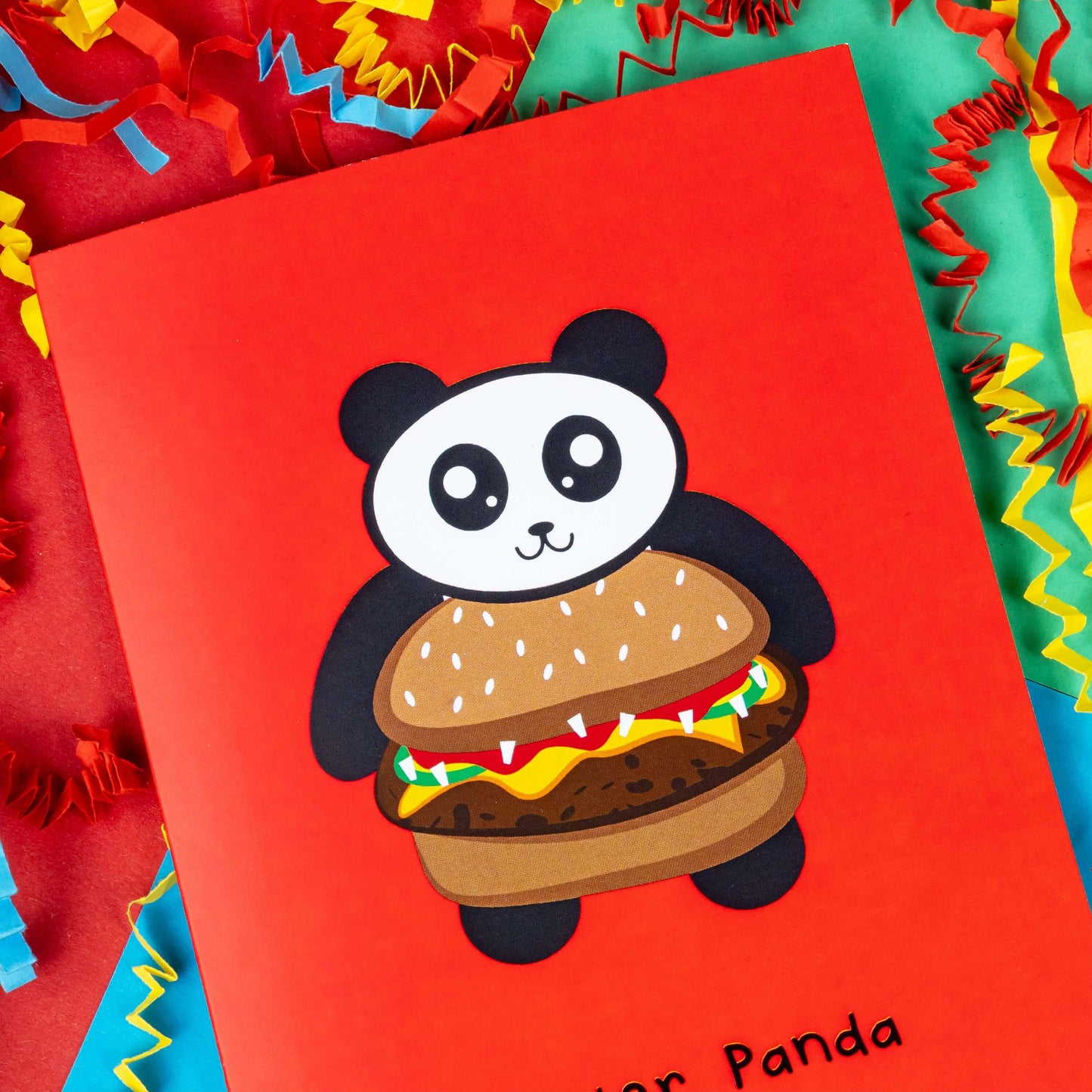 The Quarter Panda Burger Card on a red, blue and green background with yellow, red and blue crinkle card confetti. The red a6 greeting card has a smiling panda with its body being a seeded bun burger. Underneath in black reads 'quarter panda'.