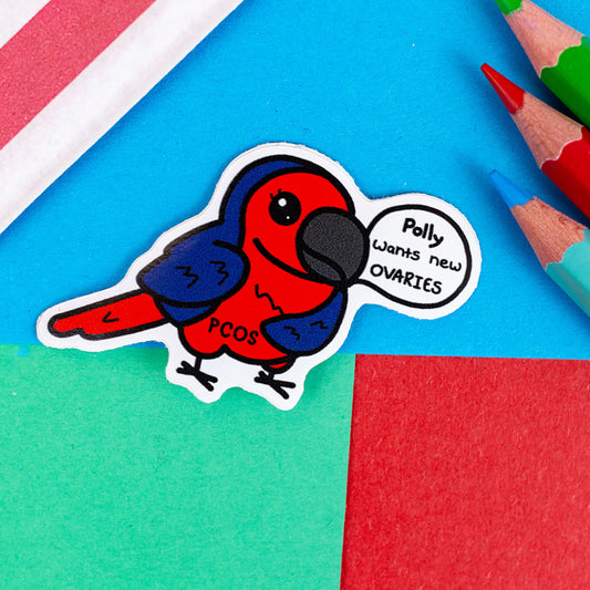 The Pollycystic Ovary Syndrome Parrot Sticker - Polycystic Ovary Syndrome PCOS on a red, blue and green background with colouring pencils and red stripe candy bag. The blue and red smiling parrot has a speech bubble reading 'polly wants new ovaries' with 'PCOS' written across its belly. The hand drawn design is raising awareness for Polycystic Ovary Syndrome PCOS.