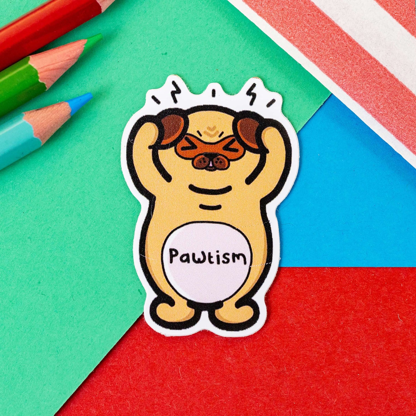 The Pawtism Dog Sticker - Autism on a red, blue and green background with colouring pencils and red stripe candy bag. The brown stressed pug dog sticker is stood up clutching its paws over its ears with black movement lines coming from its head, across its middle in black reads 'pawtism'. The hand drawn design is raising awareness for autism and neurodivergence.