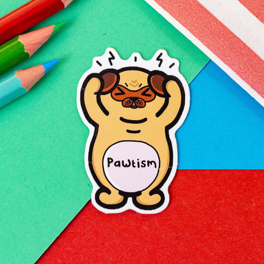 The Pawtism Dog Sticker - Autism on a red, blue and green background with colouring pencils and red stripe candy bag. The brown stressed pug dog sticker is stood up clutching its paws over its ears with black movement lines coming from its head, across its middle in black reads 'pawtism'. The hand drawn design is raising awareness for autism and neurodivergence.