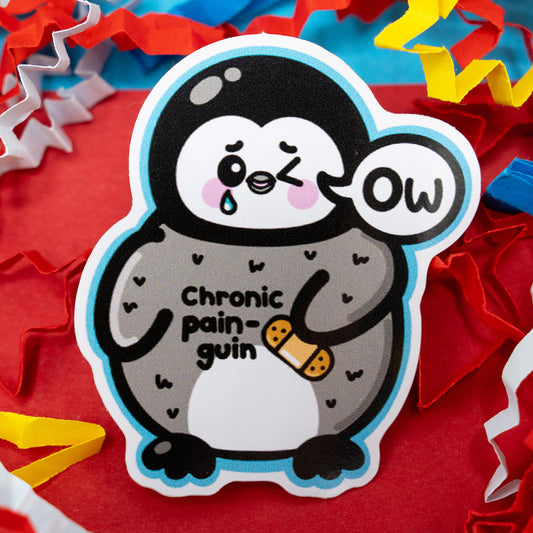 Chronic Pain-guin vinyl sticker by Innabox featuring a cute grey and white penguin holding its flipper with a bandage and a speech bubble saying “Ow.” Surrounded by red, yellow, and blue paper confetti. Glossy waterproof sticker designed to raise awareness for chronic pain and hidden disabilities in a kind and relatable way.