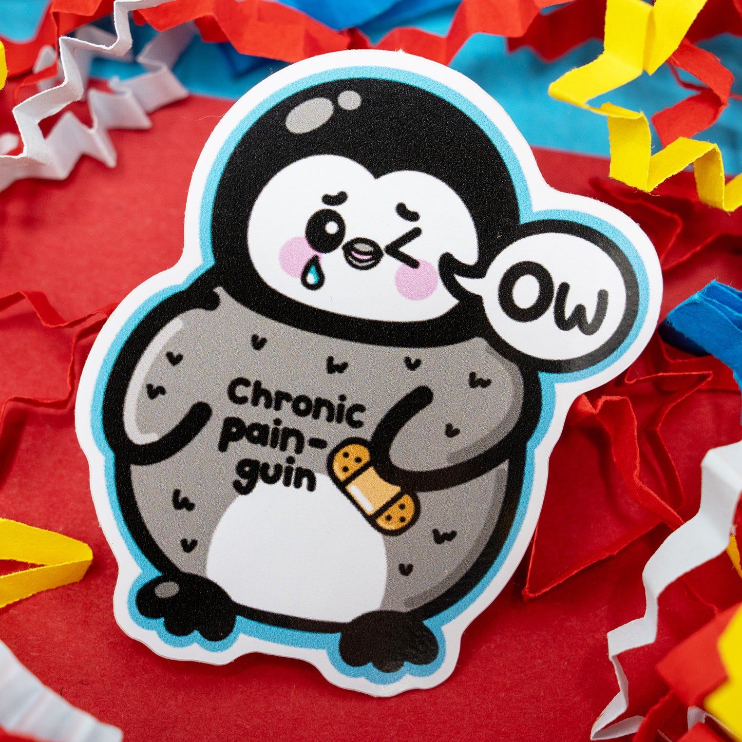 Close-up of Chronic Pain-guin vinyl sticker by Innabox showing an adorable penguin with rosy cheeks, a tear, and the text “Chronic pain-guin.” Surrounded by colourful confetti on a red background. A glossy awareness sticker spreading kindness and understanding for chronic pain and invisible illness.