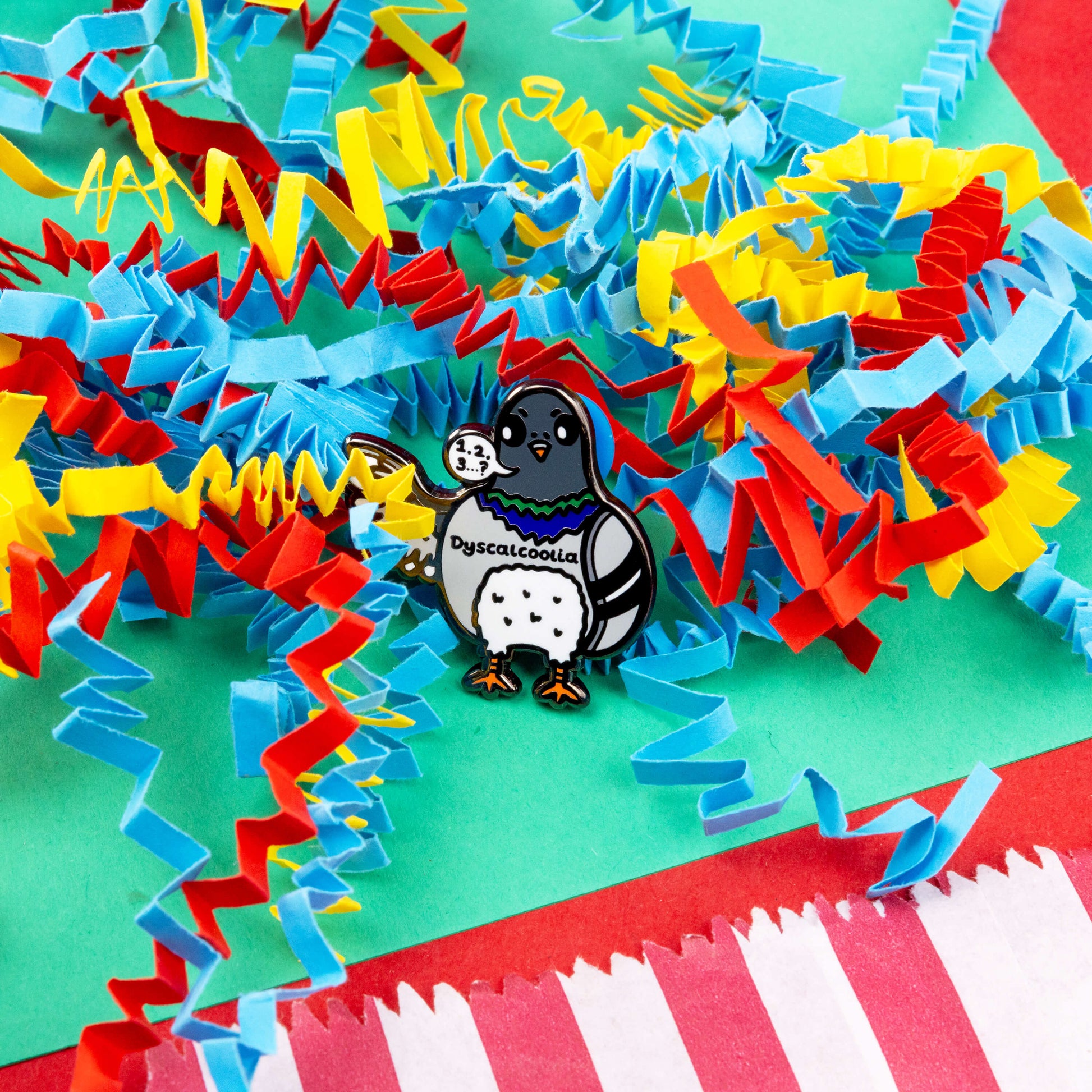 The Dyscalcoolia Pigeon Enamel Pin - Dyscalculia on a red, blue, green and yellow crinkle card confetti background. A Pigeon looking confused holding up a wing with a speech bubble full of numbers and a question mark across its chest reads 'dyscalcoolia'. The enamel pin is raising awareness for dyscalculia.