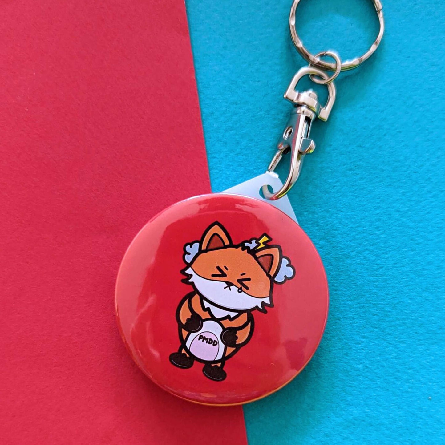 The PMDD Fox Keyring - Premenstrual Dysphoric Disorder PMDD on a red and blue background. The silver lobster clip red plastic circular keyring features a stressed fox with scrunched closed eyes and grey storm clouds above its head, across its pink belly is black text reading 'PMDD'. The hand made design is raising awareness for Premenstrual Dysphoric Disorder PMDD.
