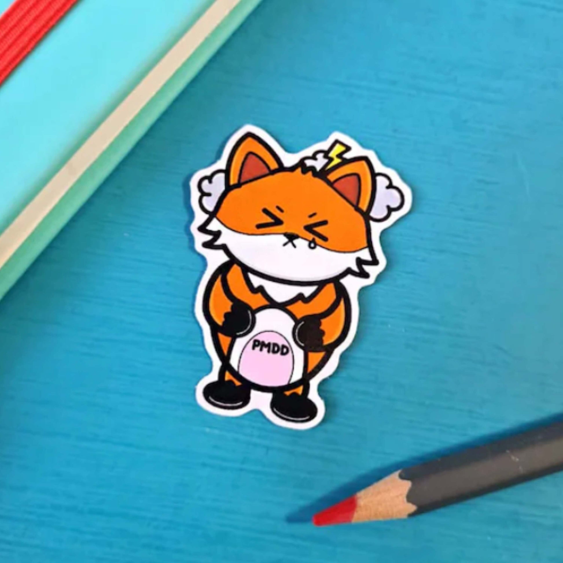 The PMDD Fox Sticker - Premenstrual Dysphoric Disorder PMDD on a blue background with a blue notebook and colouring pencil. The orange fox sticker has its eyes scrunched shut with storm clouds above its head, clutching its pink belly with black text reading 'PMDD'. The hand drawn design is raising awareness for Premenstrual Dysphoric Disorder.